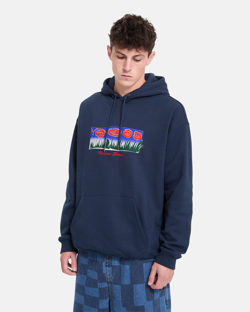 A young person wearing a navy blue Volcom Men’s Featured Artist Antonio Carrau Pullover hoodie with a colorful graphic, paired with patterned blue jeans, stands against a plain white background.