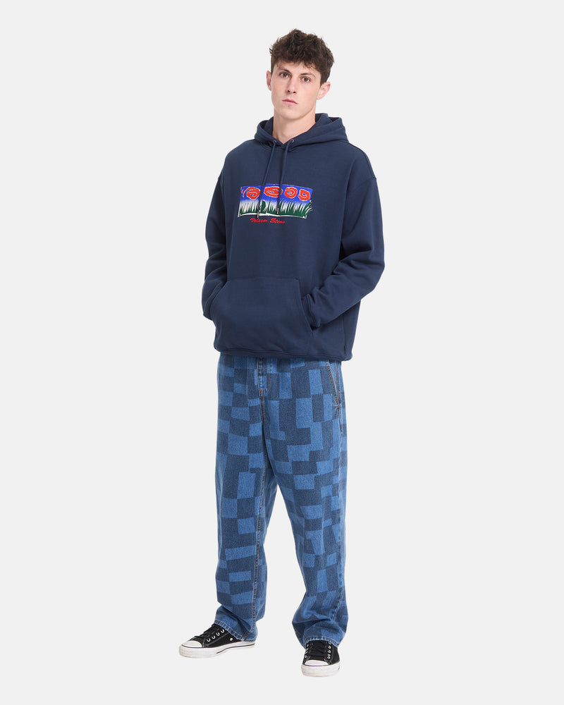 A young man stands against a plain white background wearing the Volcom Men’s Featured Artist Antonio Carrau Pullover with colorful graphics, paired with blue checkered jeans and black sneakers.