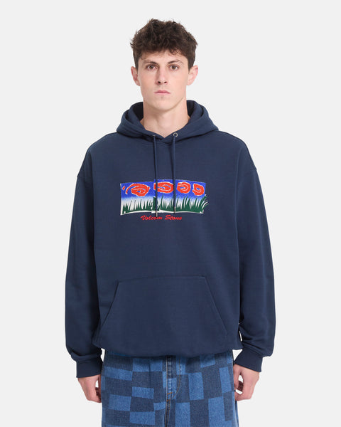 A person wearing the Men’s Featured Artist Antonio Carrau Pullover by Volcom—a navy blue heavyweight fleece hoodie adorned with a colorful graphic and text—stands against a plain white background. This Volcom hoodie includes a front pocket and is paired with blue checkered jeans.