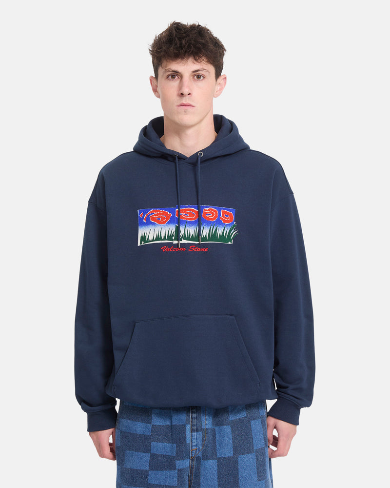 A person wearing the Men’s Featured Artist Antonio Carrau Pullover by Volcom—a navy blue heavyweight fleece hoodie adorned with a colorful graphic and text—stands against a plain white background. This Volcom hoodie includes a front pocket and is paired with blue checkered jeans.
