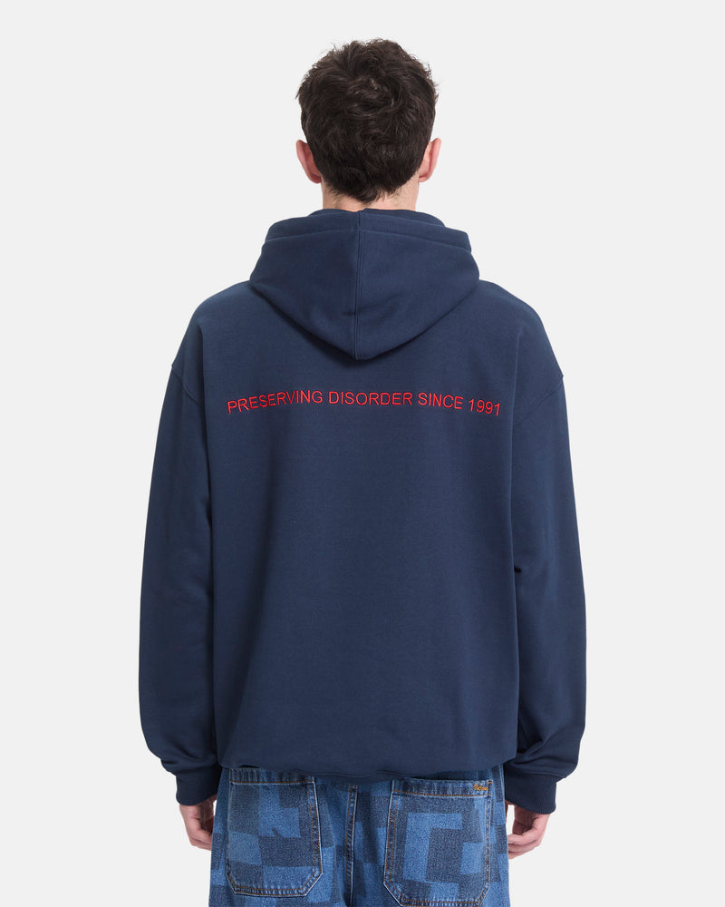 A man stands against a plain white background wearing the Volcom Men’s Featured Artist Antonio Carrau Pullover, a navy blue heavyweight fleece hoodie featuring “PRESERVING DISORDER SINCE 1991” in red text on the back.