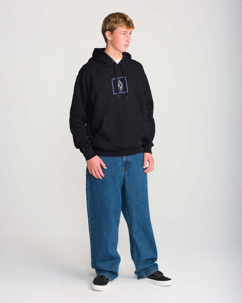 A person stands in a studio wearing a black Men’s Quadratic Pullover by Volcom with a geometric logo, loose blue jeans, and black sneakers against a plain light gray background.