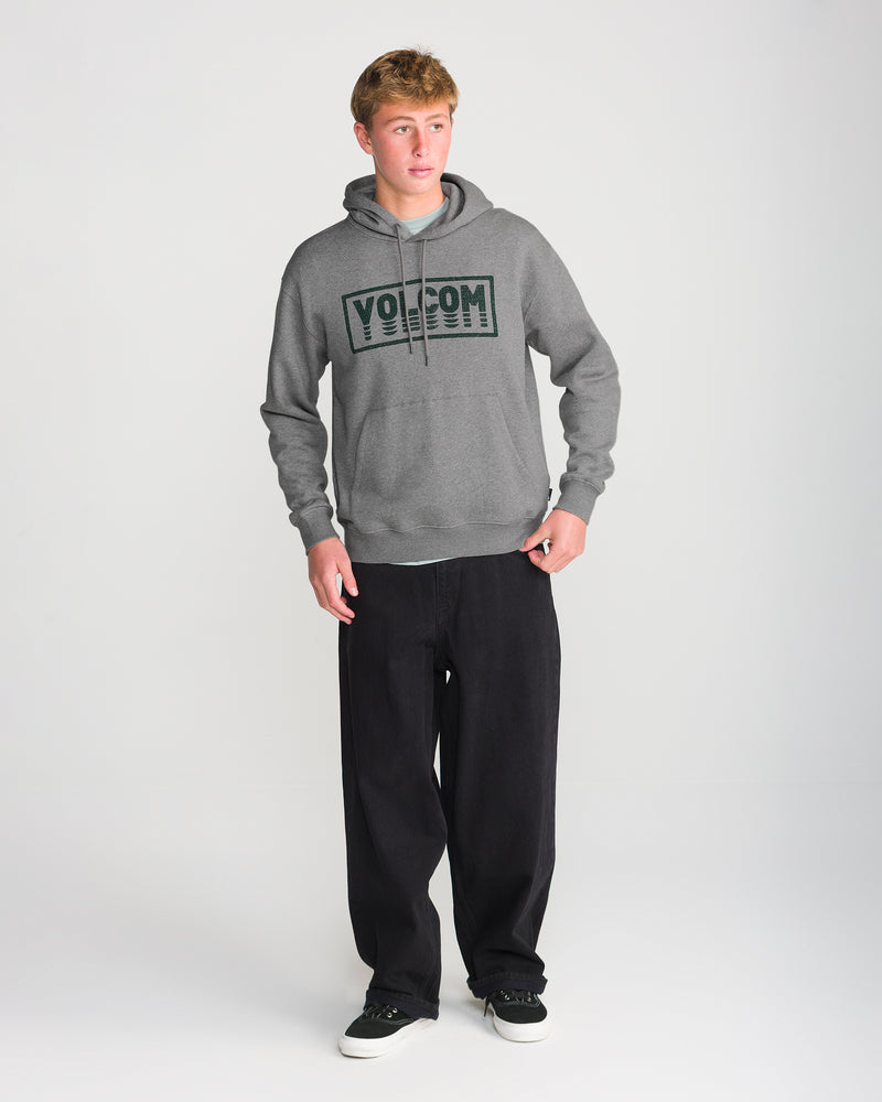 A person stands on a white background wearing the Volcom Men’s Quadratic Pullover in gray with recycled fibers, black wide-leg pants, and black shoes, looking slightly to the side with one hand on their hip.