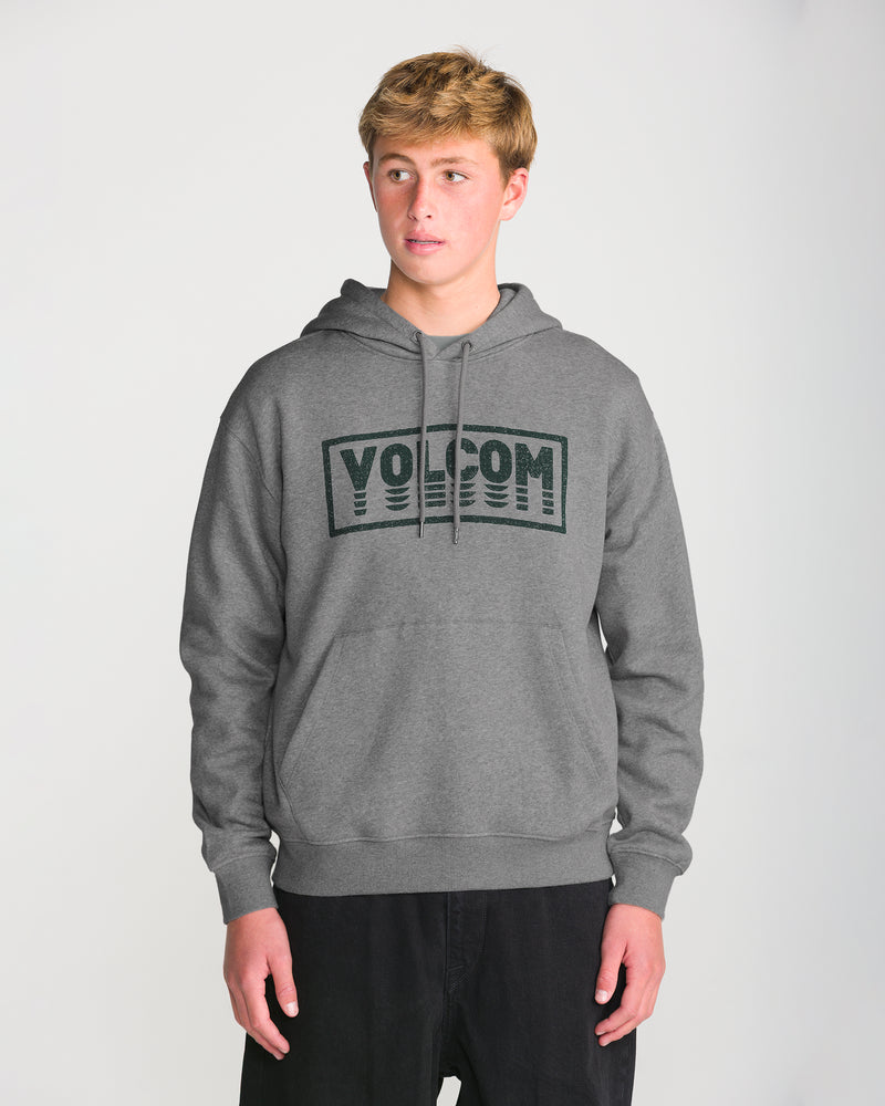 A young person in a gray Men’s Quadratic Pullover by Volcom, made with recycled fibers, and black pants stands against a white background, gazing to the side with a neutral expression.