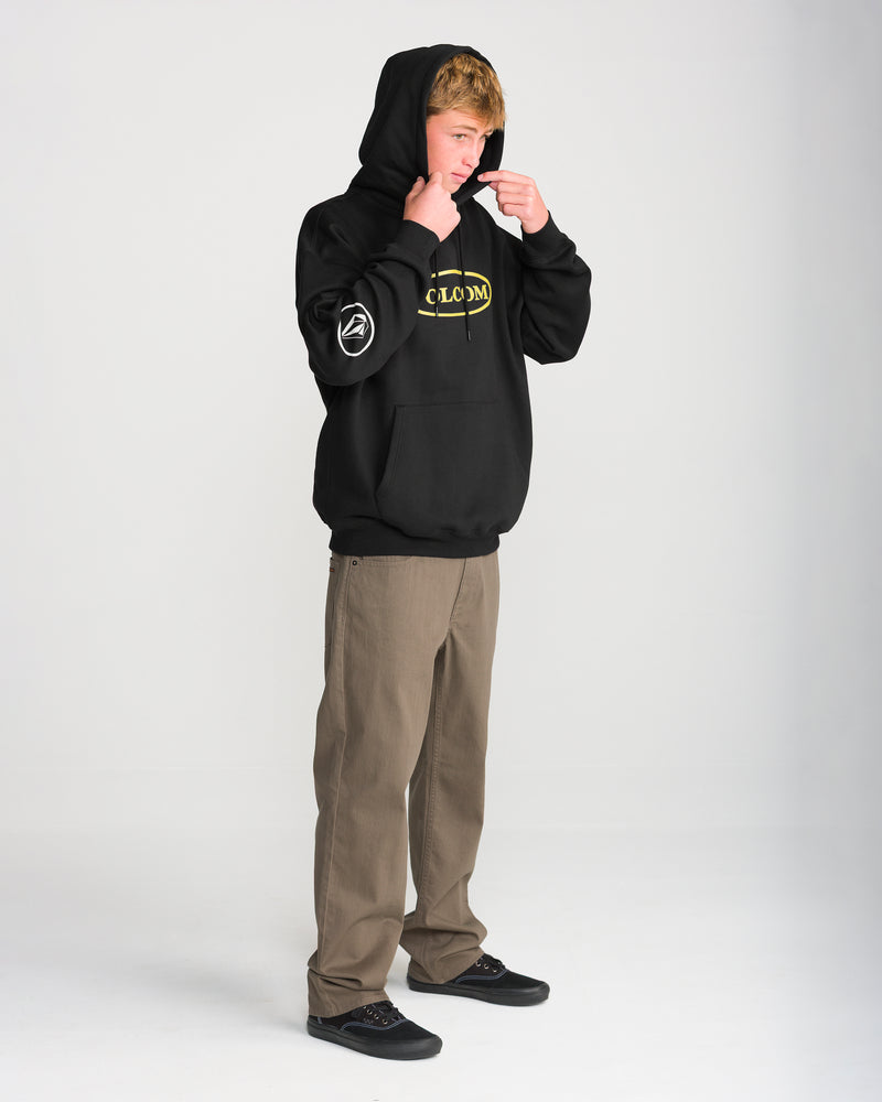 A person wears a black Volcom Men’s Remastered Stone Pullover hoodie with a yellow logo on the chest and a white graphic on the sleeve, paired with brown pants and black sneakers.