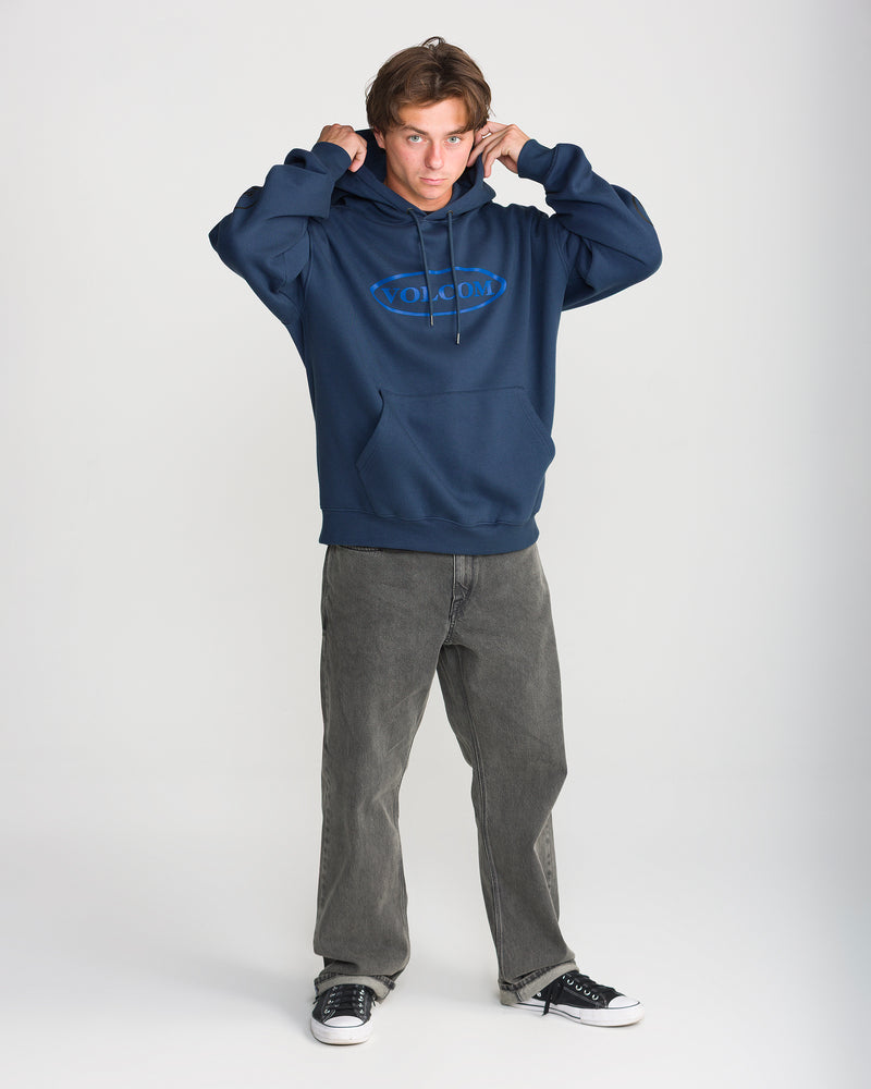 A person wearing a navy blue Men’s Remastered Stone Pullover with a blue oval logo on the chest, gray jeans, and black sneakers.