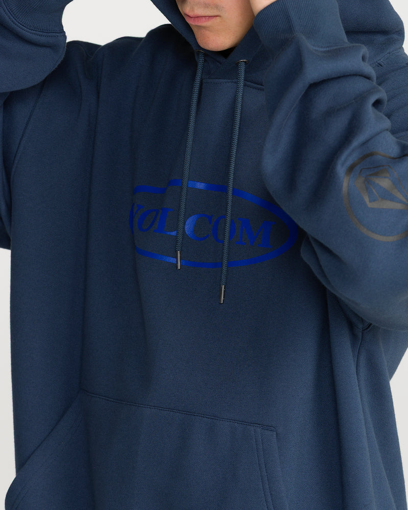 A person wearing a navy blue Volcom Men’s Remastered Stone Pullover hooded sweatshirt made of brushed-back fleece, featuring a blue oval logo graphic on the chest and a diamond emblem on the sleeve.