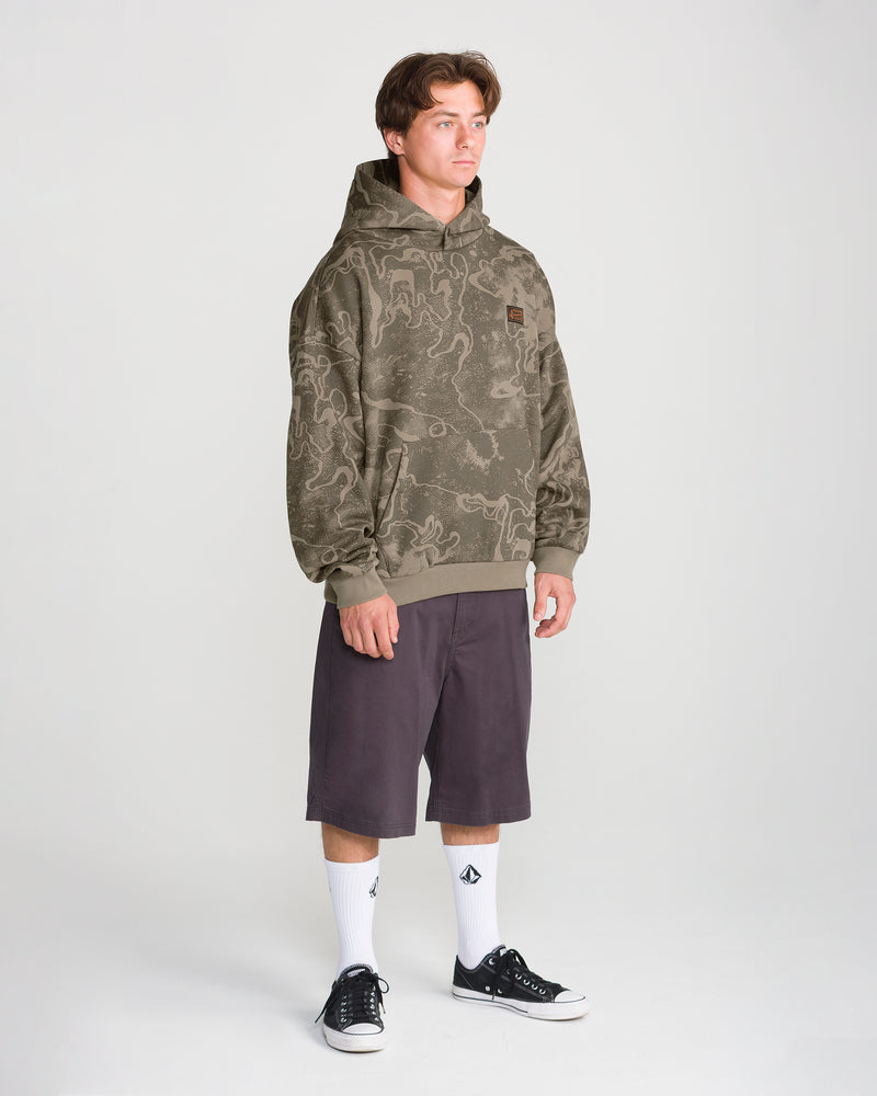A person wearing a Volcom Men’s Hailstorm Workwear Pullover in olive green with a dark brown topographical pattern, dark shorts, white socks, and black sneakers.