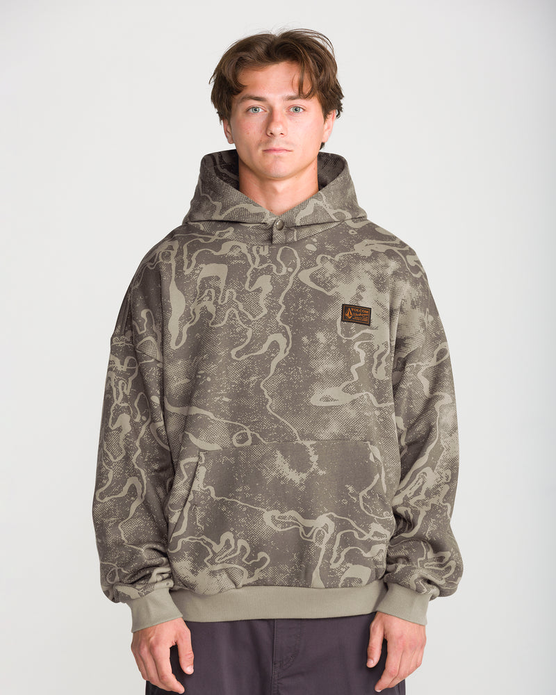 A man wears a Volcom Men’s Hailstorm Workwear Pullover, a grey camo hooded sweatshirt with a dark green marbled pattern and a square logo patch on the chest.