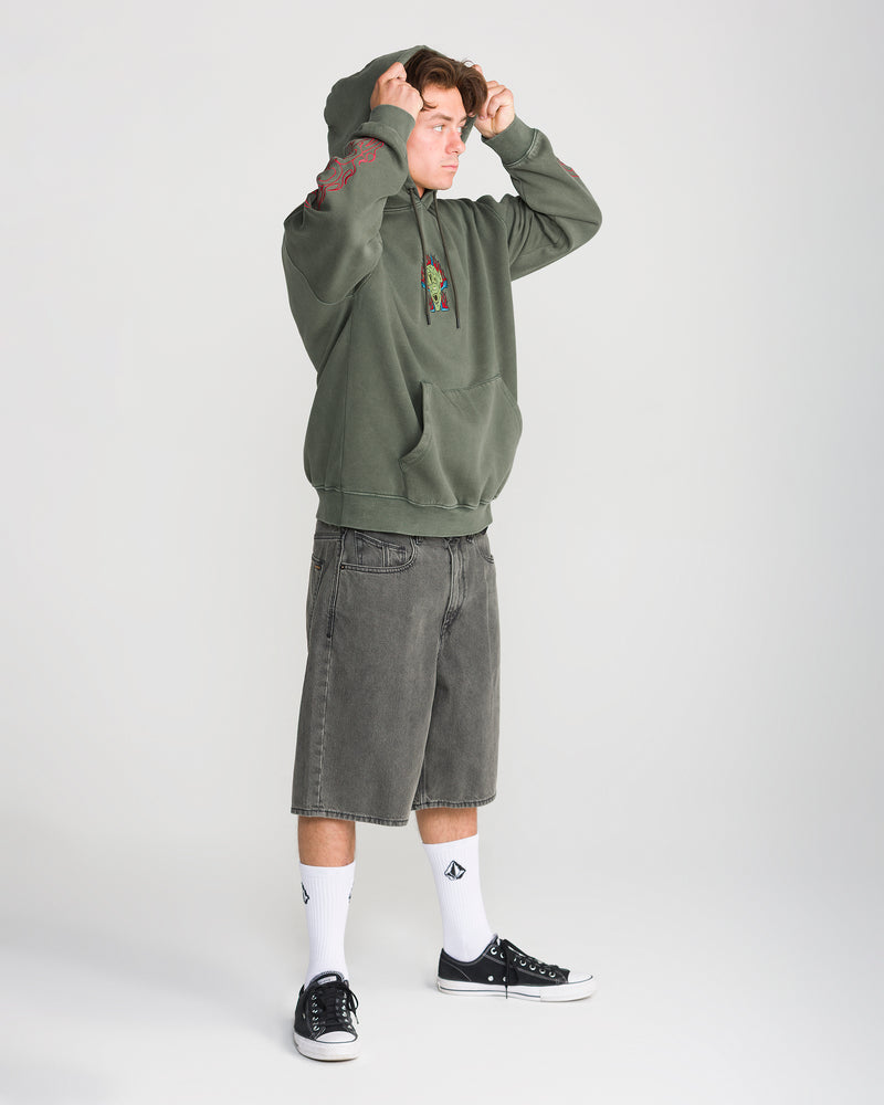 A man wears an olive green Men’s Featured Artist Casey Jones Pullover with a chest graphic and red sleeve detailing, paired with gray denim shorts, white socks, and black sneakers.