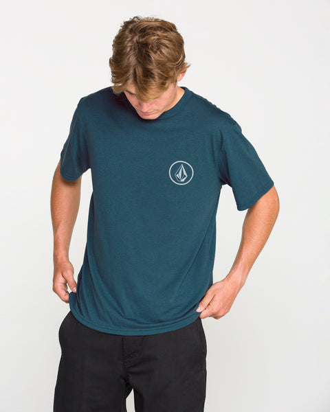A man wearing the Volcom Men's Stone Tech Short Sleeve Tee in teal, featuring a small white geometric logo on the left chest, looks down as he adjusts the hem. Crafted from quick-dry fabric with a modern fit, the T-shirt is styled with black pants against a plain white background.