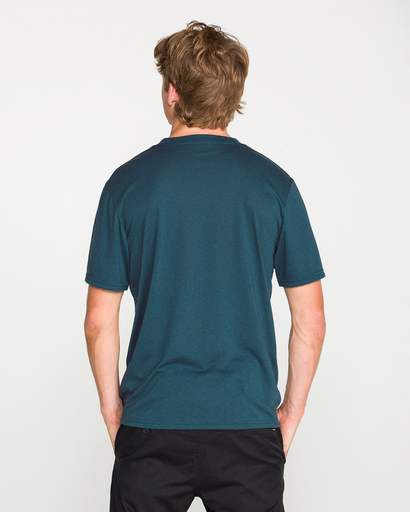 A person with light brown hair is standing with their back to the camera, wearing the Volcom Men's Stone Tech Short Sleeve Tee in teal—crafted from quick-dry fabric—paired with black pants, against a plain white background.