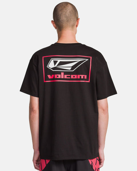 A person with a shaved head, seen from behind, wears a black Volcom Men's Surf Vitals Modstone Short Sleeve Tee with a large white and red logo and text on the back, paired with black and red shorts.