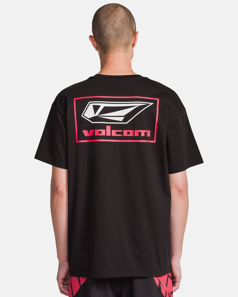 A person with a shaved head, seen from behind, wears a black Volcom Men's Surf Vitals Modstone Short Sleeve Tee with a large white and red logo and text on the back, paired with black and red shorts.