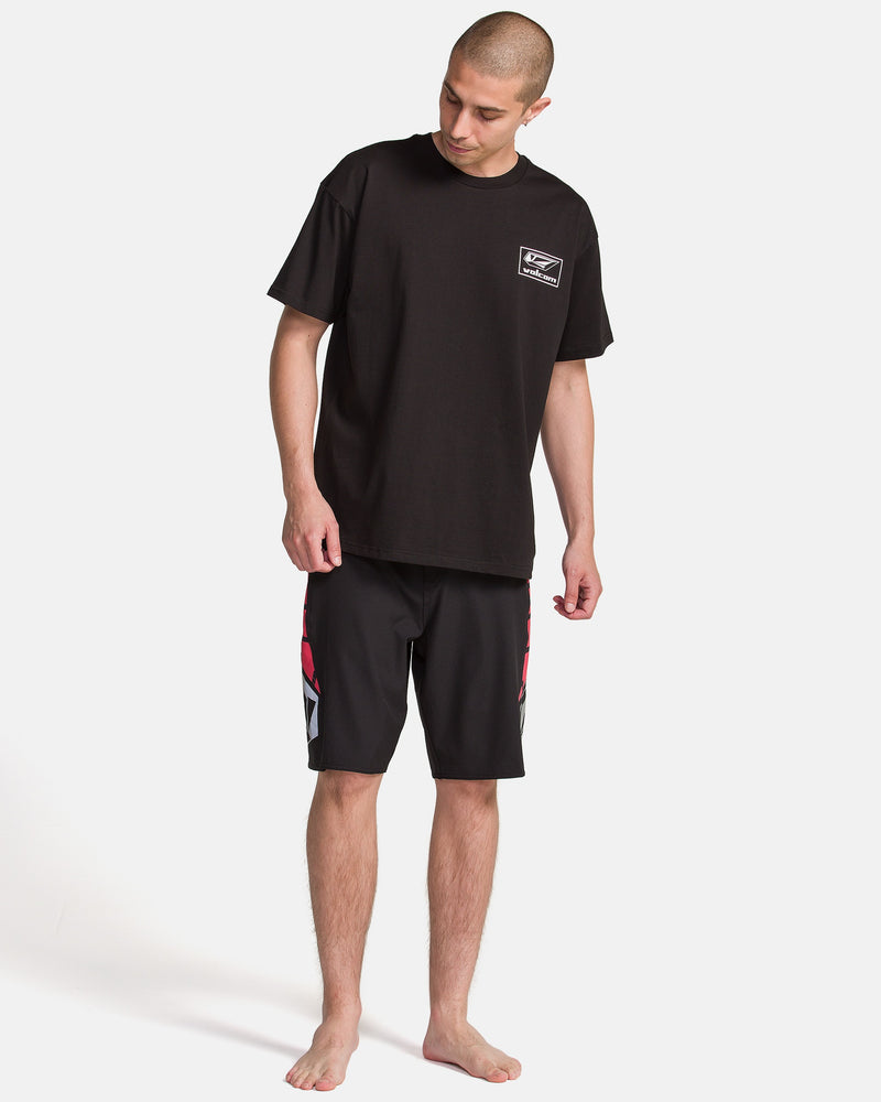 A man with short hair wears the Volcom Men's Surf Vitals Modstone Short Sleeve Tee and black knee-length shorts with red and white designs, standing barefoot on a white background and looking down to his left.