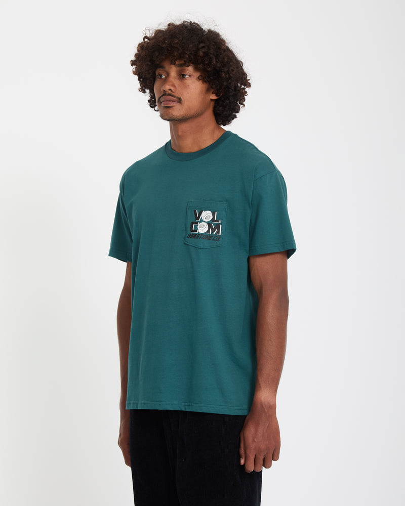 A man with curly hair wears the Volcom Men's Parted Out Pocket Short Sleeve Tee, featuring a graphic design on the chest pocket. He stands against a plain white background, looking slightly to the side.