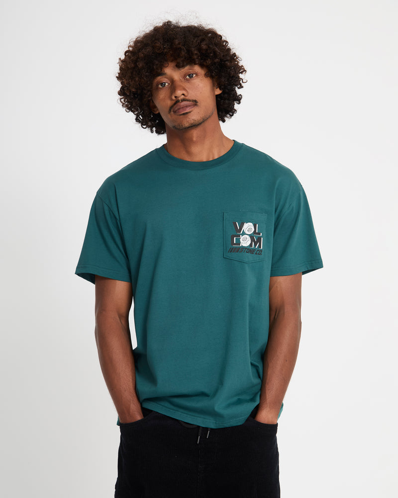 A man with curly hair wears the Volcom Men's Parted Out Pocket Short Sleeve Tee, featuring a chest pocket with the Volcom logo, standing against a white background with his hands in his pockets.