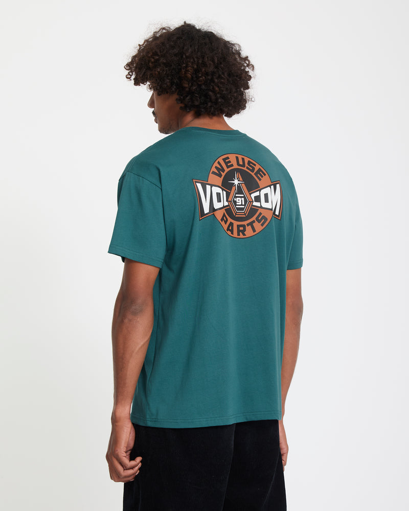 A man with curly hair, seen from behind, wears the Volcom Men's Parted Out Pocket Short Sleeve Tee in teal, featuring a large circular "WE USE VOLCOM PARTS" logo on the back in brown, orange, and white.