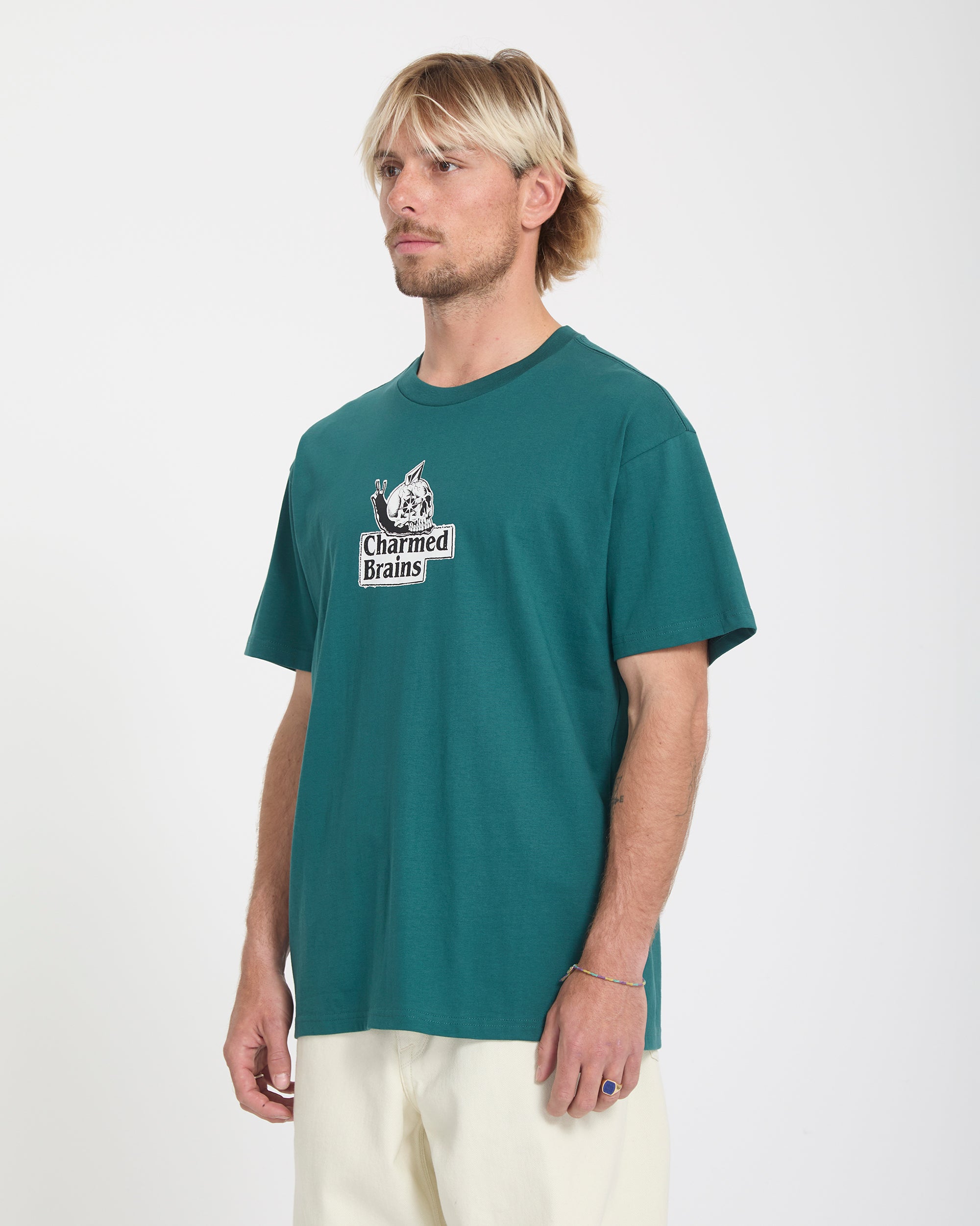 A man with blonde hair and a beard wears the Volcom Men's Charmed Brains Short Sleeve Tee in teal and light pants, standing against a plain white background.