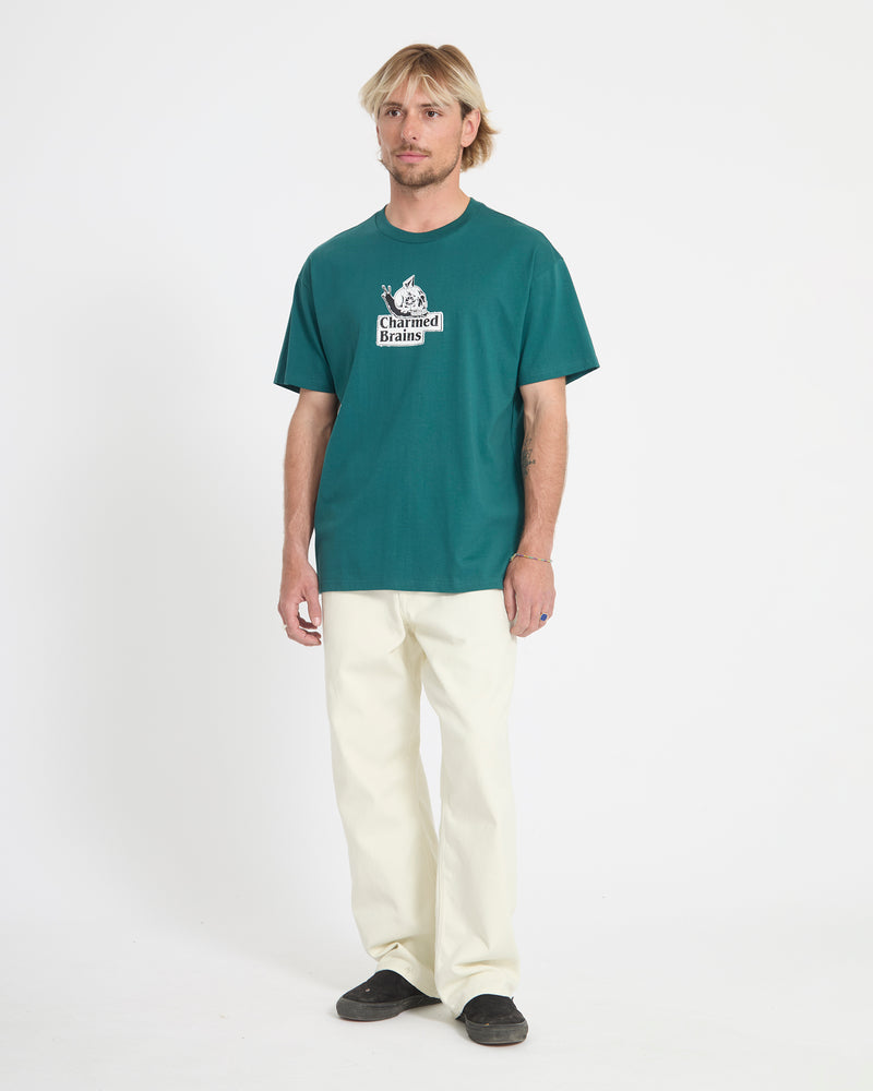A person stands against a plain white background, wearing the Volcom Men's Charmed Brains Short Sleeve Tee in teal, paired with off-white pants and black shoes. Their hands are relaxed at their sides.