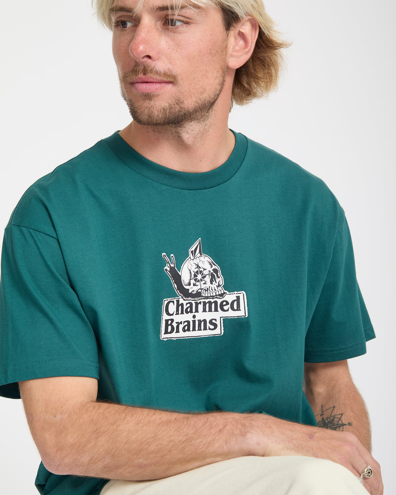 A blond man with facial hair wears the Volcom Men's Charmed Brains Short Sleeve Tee, featuring a brain and pencils graphic with the text "Charmed Brains" on the front. He sits against a plain background, looking to the side.