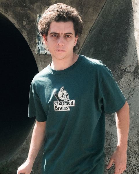 A young man with curly hair stands outside by a concrete structure, looking seriously at the camera while wearing the Volcom Men's Charmed Brains Short Sleeve Tee.