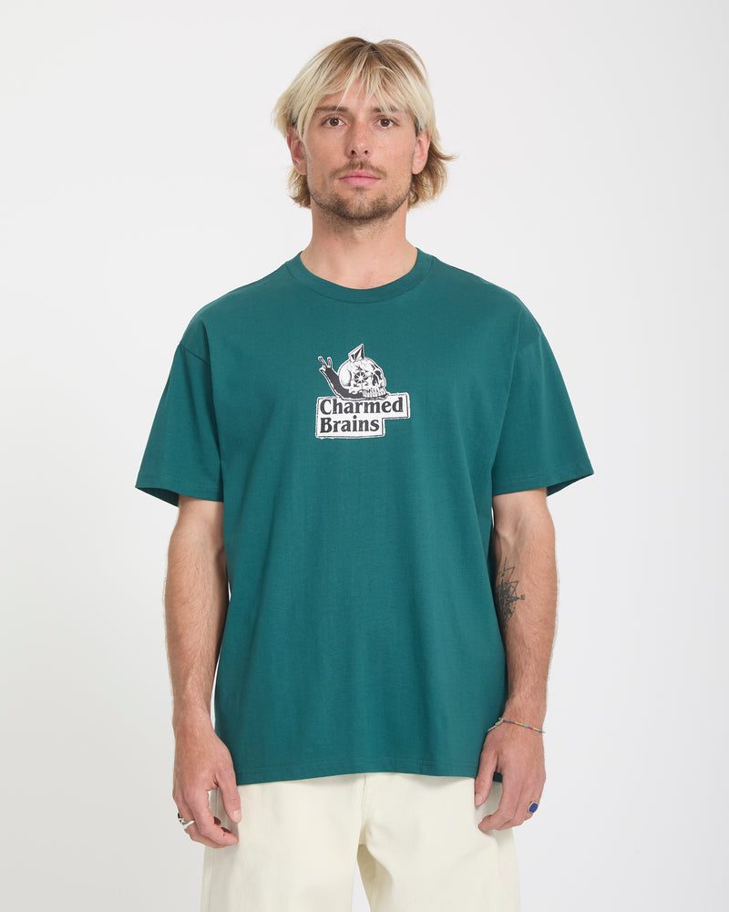 A man with blonde hair and a short beard wears the Volcom Men's Charmed Brains Short Sleeve Tee in teal and off-white pants, standing against a plain white background.