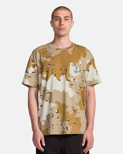 A person with short hair and a neutral expression faces forward wearing the Volcom Men's Desert Beers Pocket Short Sleeve Tee, a loose-fitting beige and brown camo shirt with an all-over print and a colorful patch on the left chest pocket.