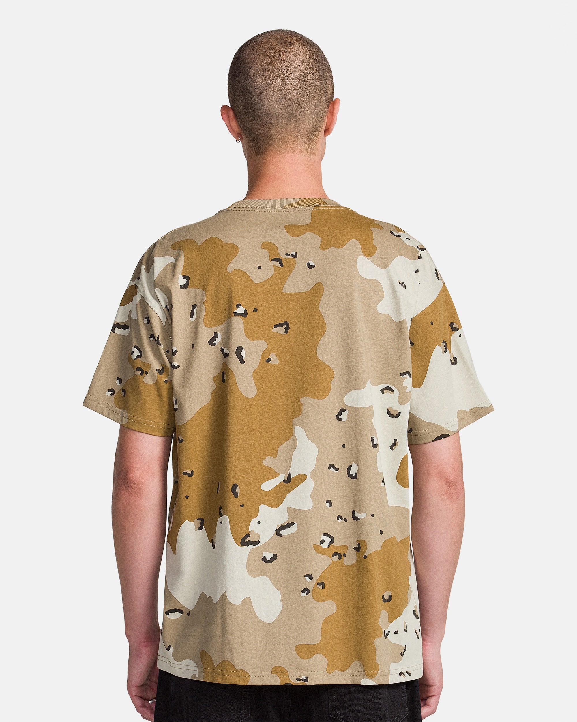 A person with a shaved head stands facing away from the camera, wearing a Volcom Men's Desert Beers Pocket Short Sleeve Tee in beige and brown camouflage, paired with black pants against a plain white background.