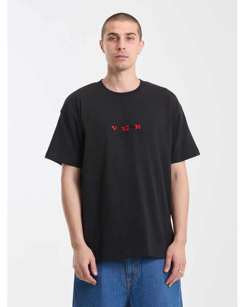 A man with short hair wears the Volcom Men's Featured Artist Seth Conboy Loose Short Sleeve Tee in black, featuring small red and white Volcom lettering on the chest, paired with blue jeans, standing against a plain white background.