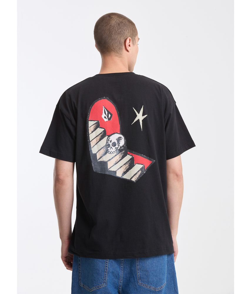 A person with short hair, wearing Volcom’s Men's Featured Artist Seth Conboy Loose Short Sleeve Tee in black—featuring a skull on a staircase, red background, and white star graphic on the back—and blue denim shorts, is facing away.