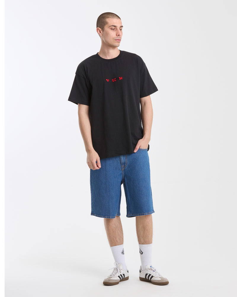 A man wears the Volcom Men's Featured Artist Seth Conboy Loose Short Sleeve Tee, styled with blue knee-length denim shorts, white socks, and sneakers. He stands against a white background with hands in pockets, looking slightly to the side.