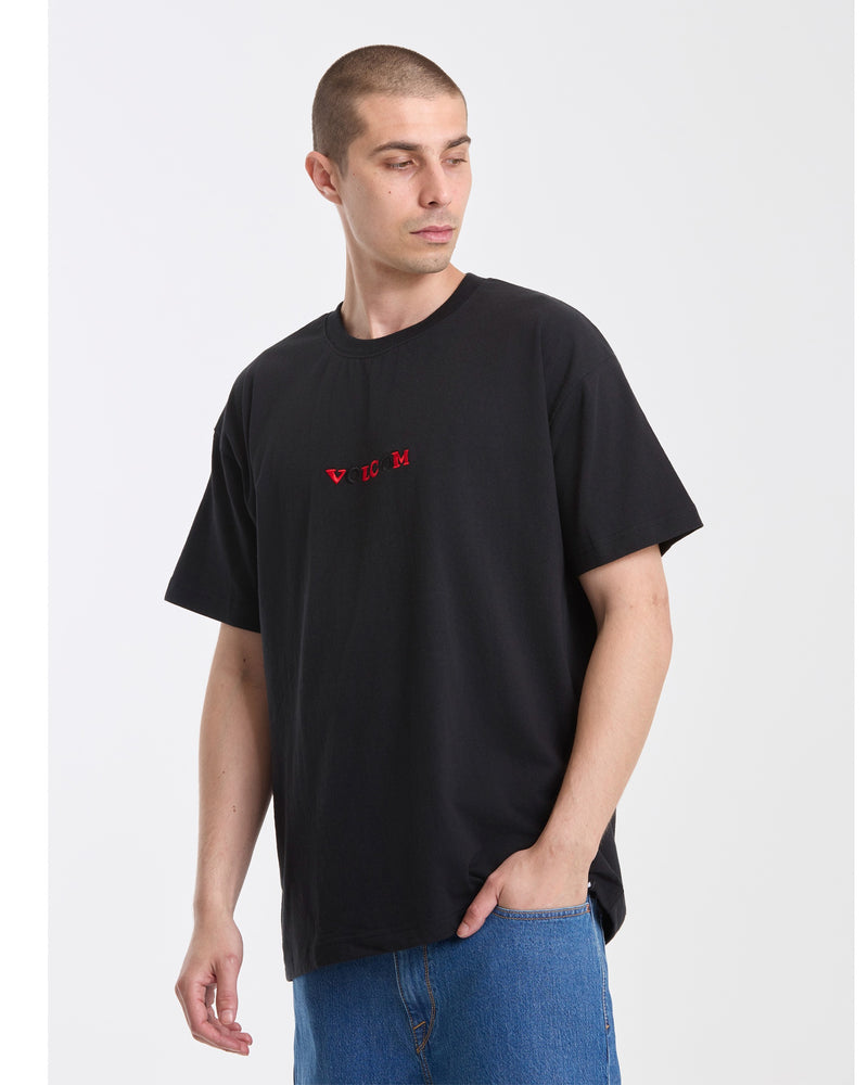 A man with short brown hair models the Volcom Men's Featured Artist Seth Conboy Loose Short Sleeve Tee, featuring small red chest text, paired with blue jeans. He stands against a plain white background, looking left with one hand in his pocket.