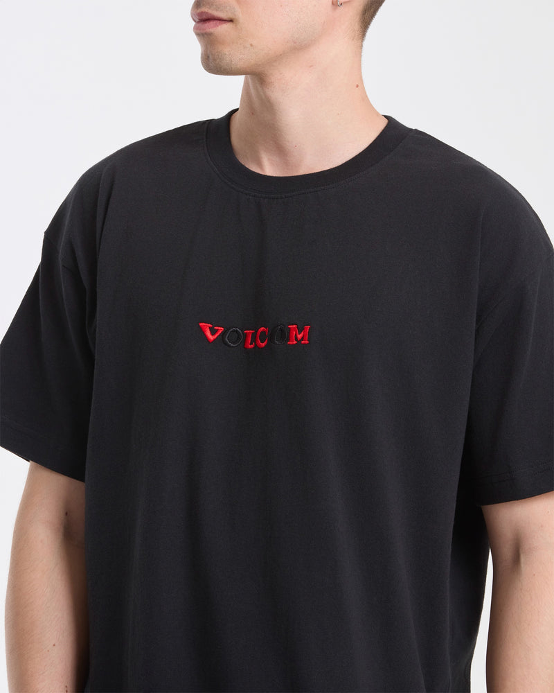 A man wears the Volcom Men's Featured Artist Seth Conboy Loose Short Sleeve Tee, a black shirt with "VOLCOM" embroidered in red and white on the chest, standing against a plain white background with his face partially out of view.