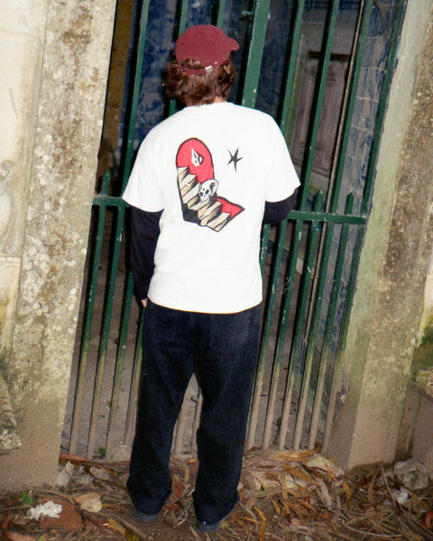 A person wears the Volcom Men's Featured Artist Seth Conboy Loose Short Sleeve Tee, featuring a red snake and skull design, while standing before a green metal gate amid old stone pillars and fallen leaves.