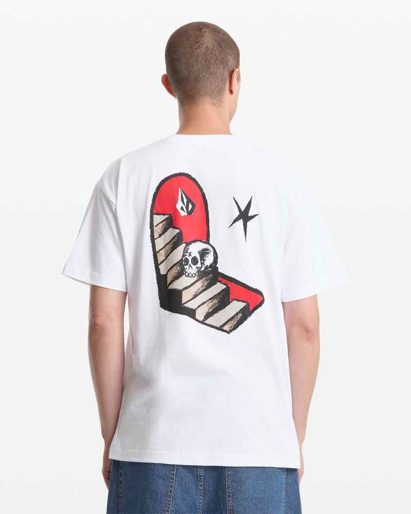 A person with short hair wears blue jeans and the Volcom Men's Featured Artist Seth Conboy Loose Short Sleeve Tee, which is white with an illustrated back design of a red and brown staircase, a skull, and a black star.