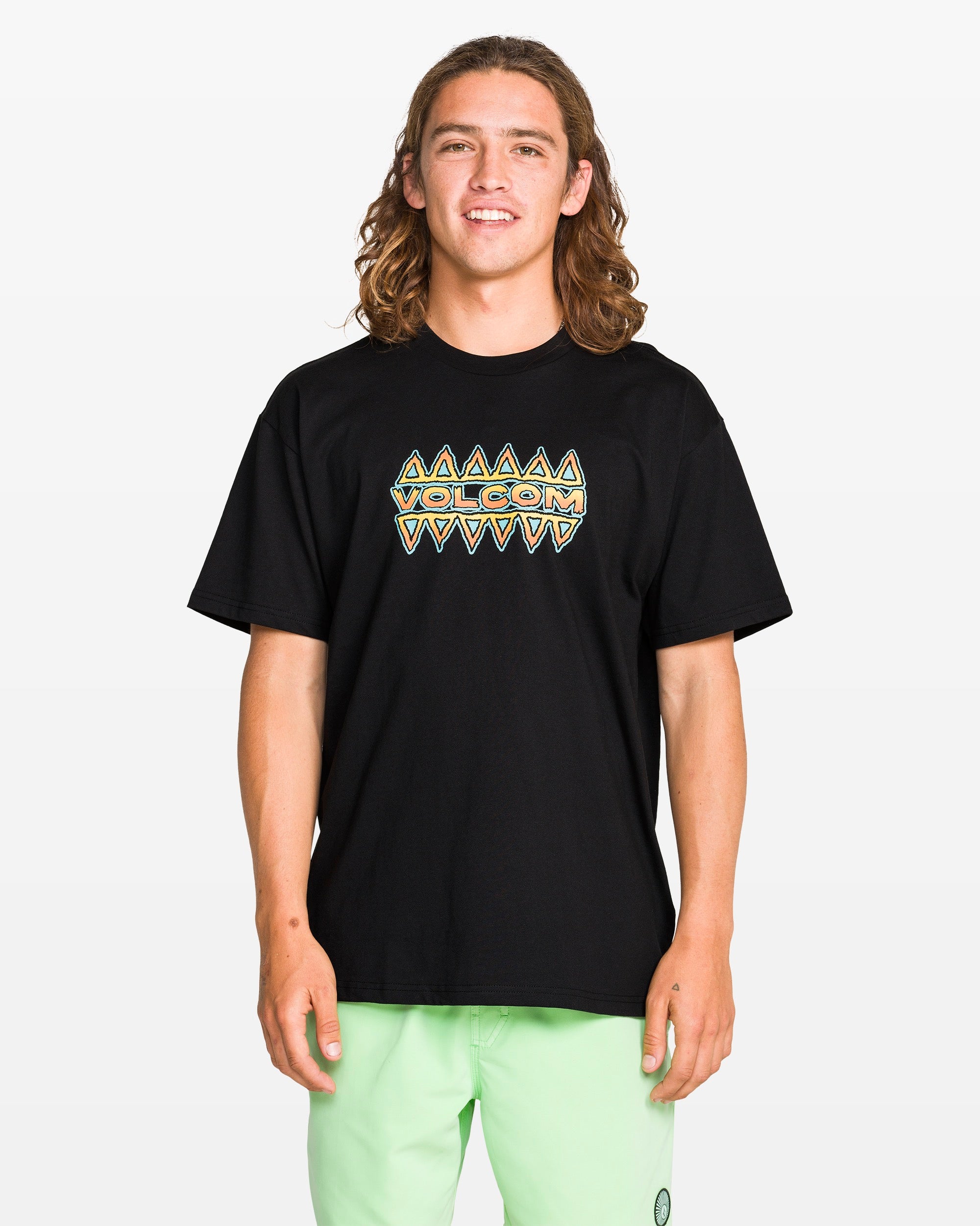 A young man with long brown hair stands and smiles against a plain white background, wearing the Volcom Men's Bodie Short Sleeve Tee featuring a colorful geometric pattern, paired with pale green shorts.