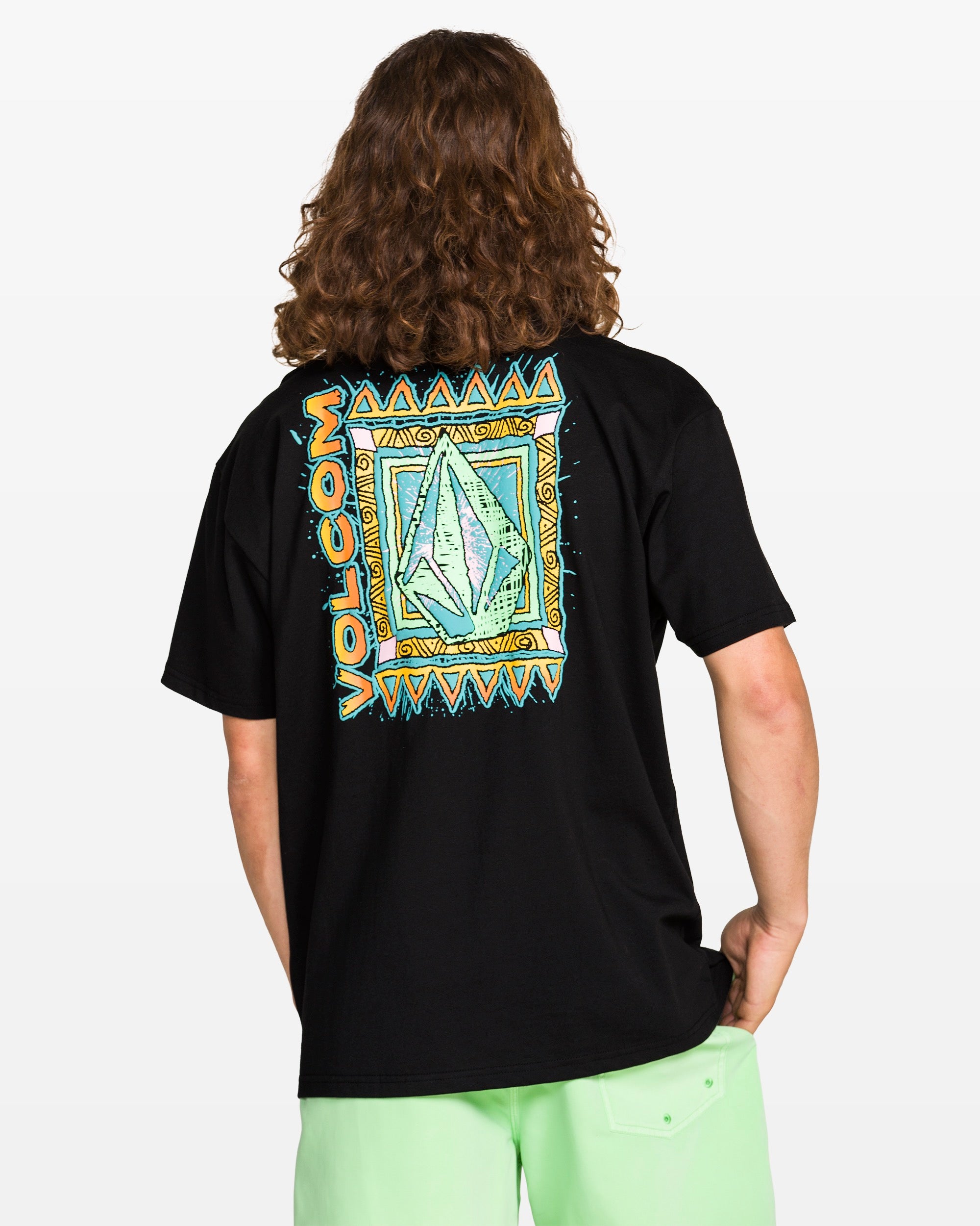 A person with long, wavy hair wears the Volcom Men's Bodie Short Sleeve Tee in black, featuring a colorful geometric logo and tribal-style border on the back, paired with light green shorts, against a plain white background.