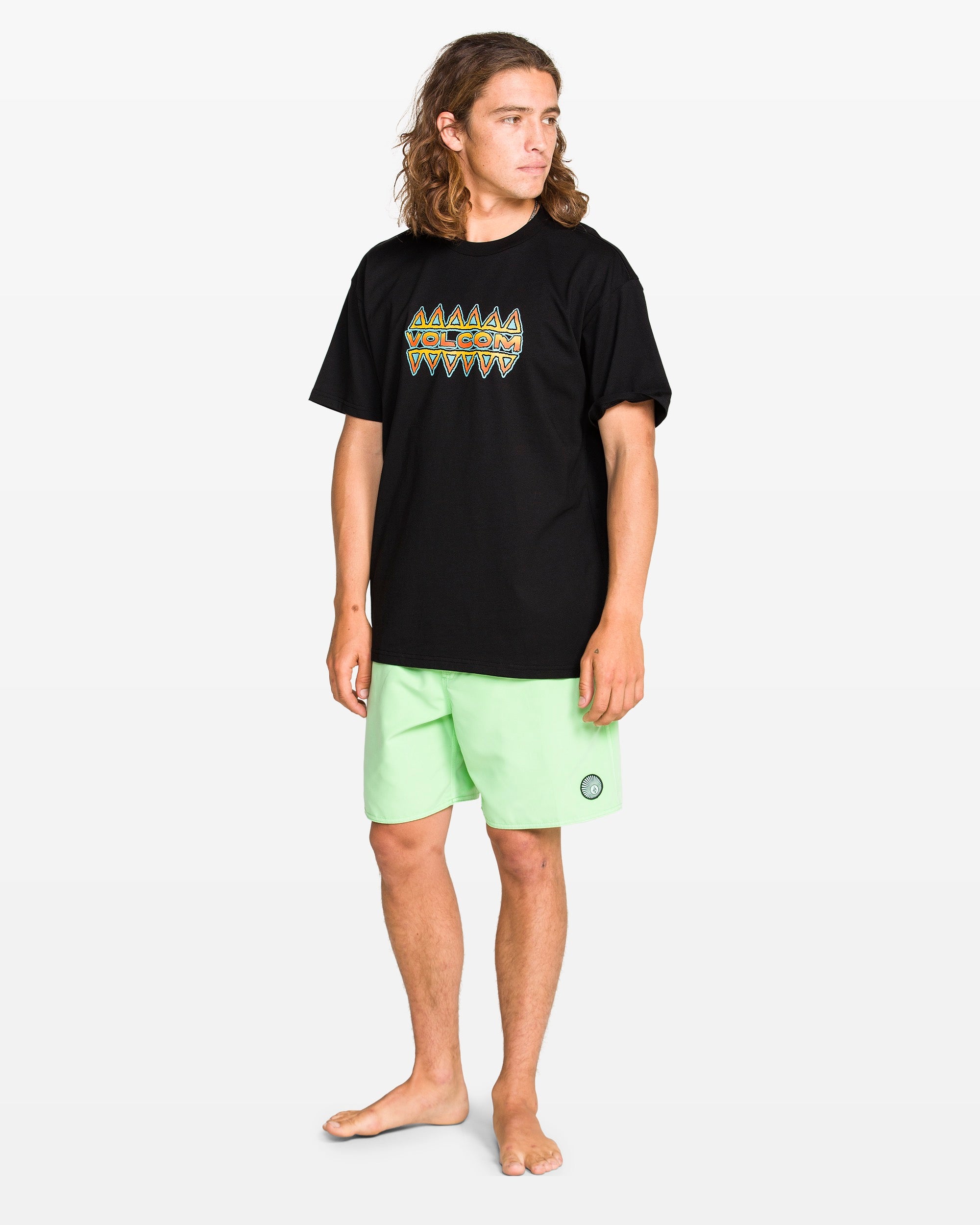 A person with long hair is wearing the Volcom Men's Bodie Short Sleeve Tee in black and light green shorts, standing barefoot on a plain white background and looking off to the side.