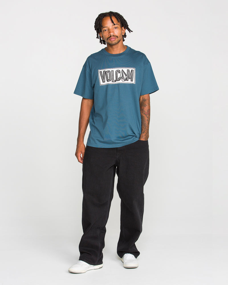 A man with short dreadlocks and a tattoo on his left forearm stands against a white background wearing the Volcom Men’s Studded Short Sleeve Tee in teal, made from premium ringspun cotton, paired with loose black pants and white shoes.