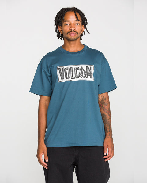 A man with short dreadlocks, dressed in black pants and a blue Volcom Men’s Studded Short Sleeve Tee featuring a soft-hand screen print, stands facing the camera against a plain white background. Crafted from premium ringspun cotton, this tee merges comfort and bold style.