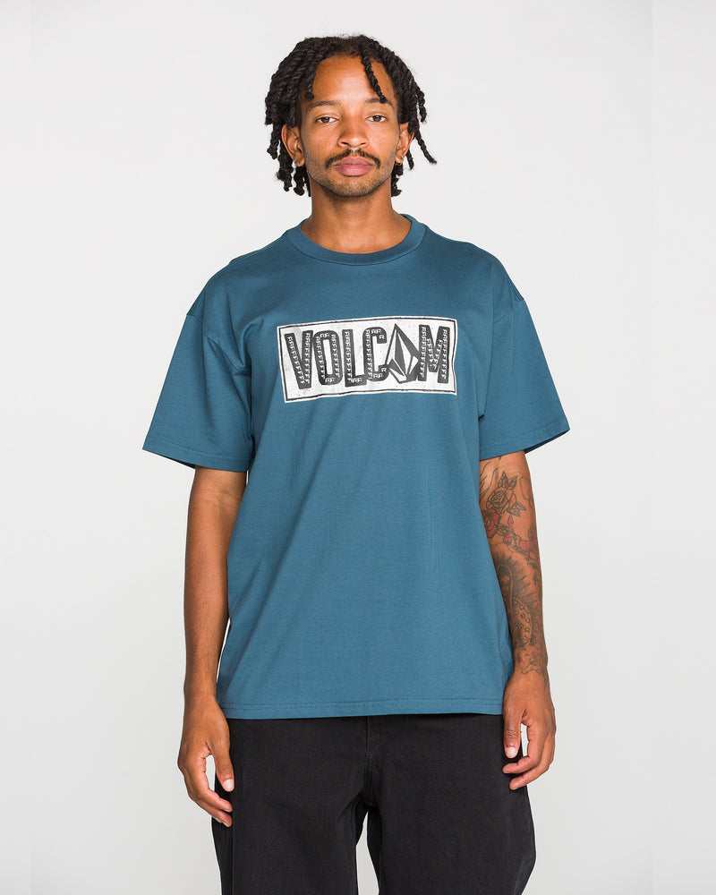 A man with short dreadlocks, dressed in black pants and a blue Volcom Men’s Studded Short Sleeve Tee featuring a soft-hand screen print, stands facing the camera against a plain white background. Crafted from premium ringspun cotton, this tee merges comfort and bold style.