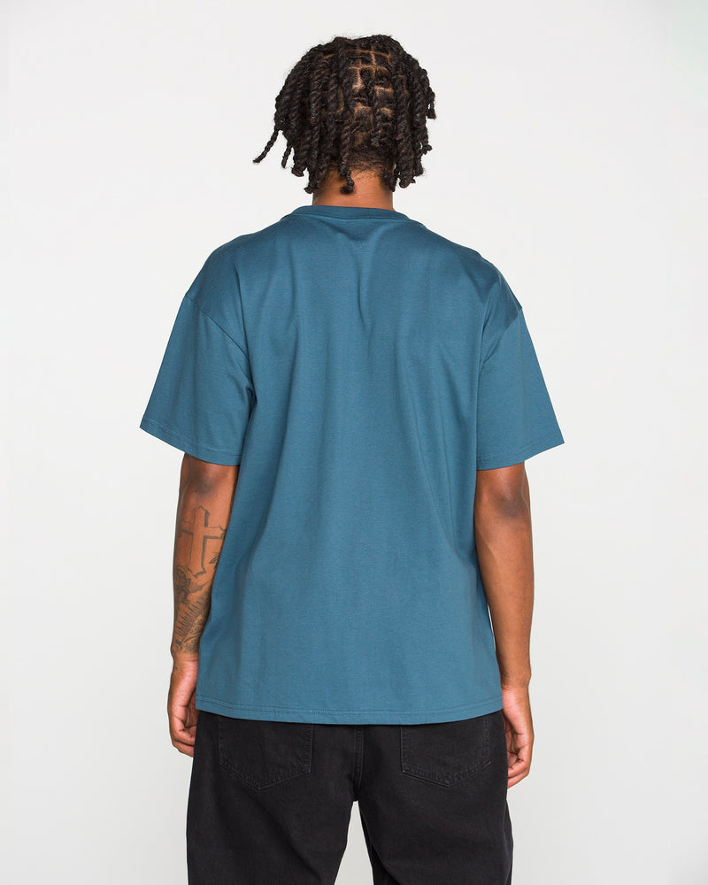 A person with short dreadlocks, dressed in a teal Volcom Men’s Studded Short Sleeve Tee made from premium ringspun cotton and black pants, stands with their back to the camera against a plain white background.