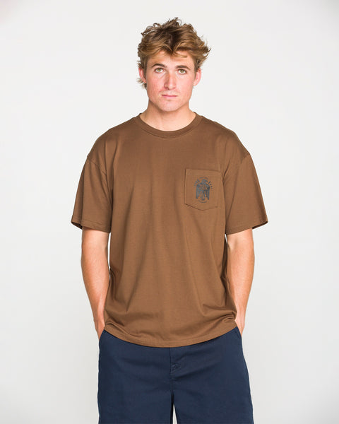 A person with short, tousled hair wears the Volcom Men's Troubador Short Sleeve Tee—a brown, loose-fit shirt made from heavyweight ringspun cotton with a small chest pocket—and pairs it with navy blue shorts while standing against a plain white background.