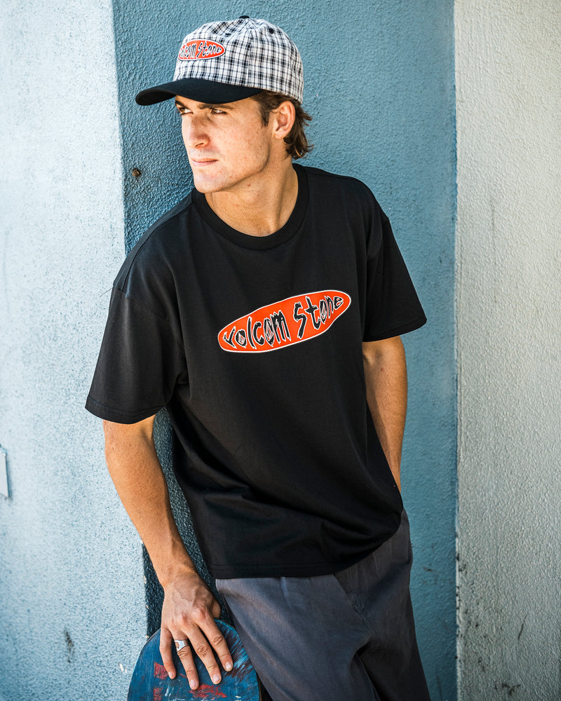A person wearing a plaid cap and the Volcom Men’s Scratch Stone Short Sleeve Tee, crafted from heavyweight ringspun cotton, leans against a blue wall holding a skateboard and looking to the side.