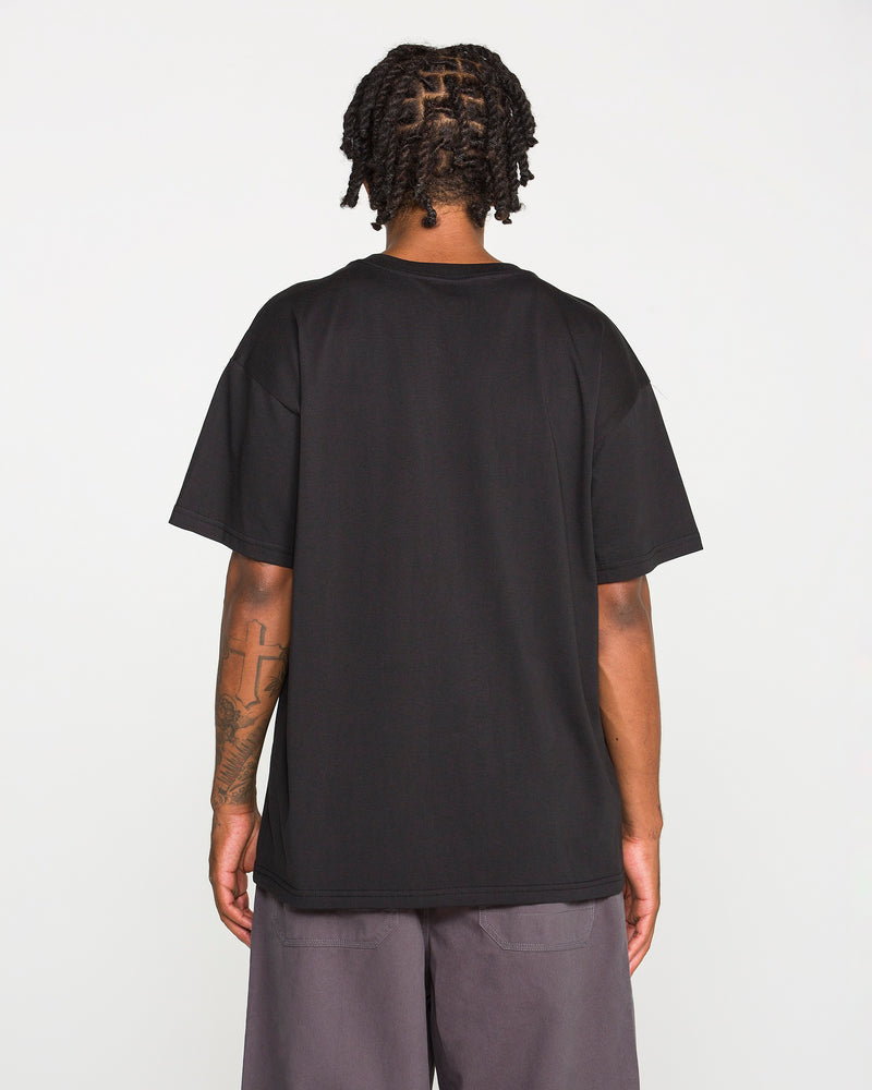 Back view of a person with dark braided hair wearing the Volcom Men's Scratch Stone Short Sleeve Tee in plain black and grey shorts. A large cross tattoo is visible on their right forearm. They are standing against a plain, light grey background.