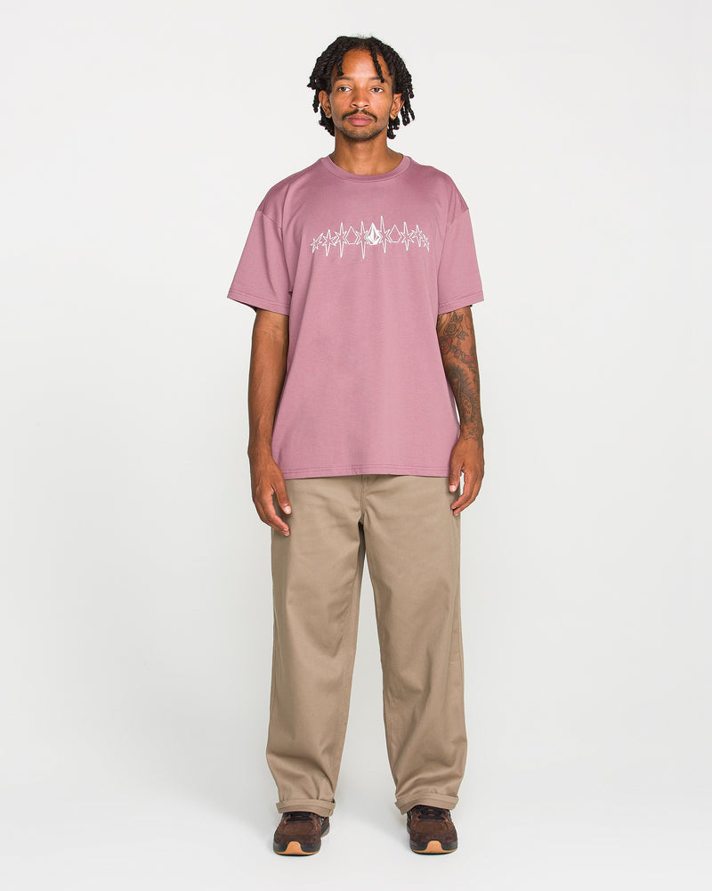 A man with short dreadlocks wears the Volcom Men's Repeater Short Sleeve Tee in a mauve hue, featuring a loose fit and crafted from heavyweight ringspun cotton. He pairs it with beige pants and brown shoes, standing facing forward against a plain white background with a neutral expression.
