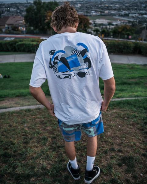 A person stands on grass overlooking a city, wearing the Volcom Men’s Featured Artist Head Check Short Sleeve Tee in white, featuring striking blue collage artwork on the back and paired with blue camo shorts. Made from premium ringspun cotton, the tee’s bold graphic is visible as they stand with their back to the camera, holding the shirt near their hips.