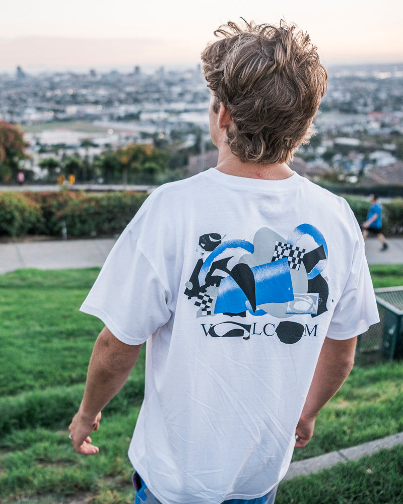 A person with tousled hair stands outdoors on grass, facing away. They wear the Volcom Men’s Featured Artist Head Check Short Sleeve Tee, made from premium ringspun cotton and featuring a striking blue, black, and white graphic collage artwork along with the word VOLCOM on the back. A cityscape is visible in the background.
