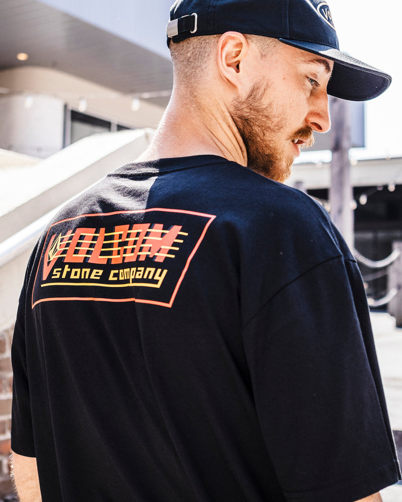 A man in a black cap and Volcom Men's Round Out Short Sleeve Tee, featuring a colorful graphic on the back, stands outdoors near stairs facing slightly away from the camera.